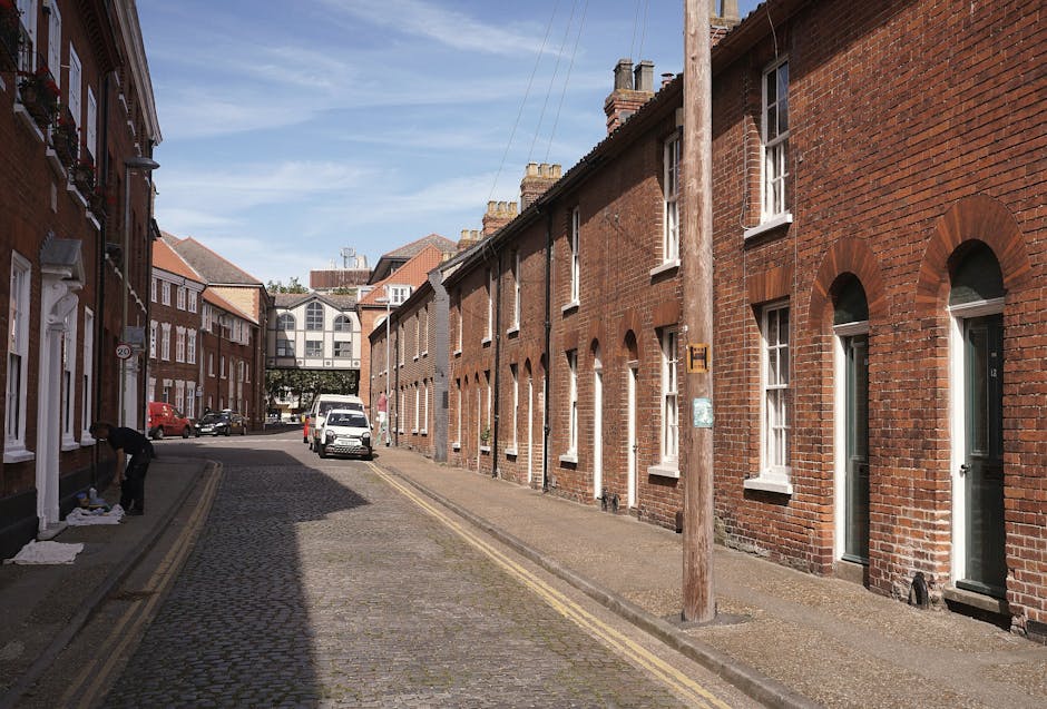 In front of a traditional brick building housing a bar and restaurant, there is a black commercial waste bin labeled 'COMMERCIAL WASTE ONLY' positioned on the pavement. The bin appears to be made of metal with a reflective surface, partially filled with flattened cardboard boxes and other waste materials, some sticking out over the top. It is located immediately outside the establishment, which has large glass windows framed in dark red wood paneling, and a sign indicating food and drink offerings. To the left of the bin, a small blackboard sign advertises a self-service buffet, while to the right, the pavement is partly obstructed by a short black bollard and a concrete street pole supporting a street sign reading 'CRISPIN STREET.' Behind the bin, the street features a painted bicycle lane marking and a parked delivery truck in the background, as well as bicycles secured to a bike rack further along the sidewalk. The scene reflects a typical urban setting where private waste collection may be used for commercial premises as part of alternative waste handling arrangements, aligned with rubbish removal services in Kingston upon Thames.