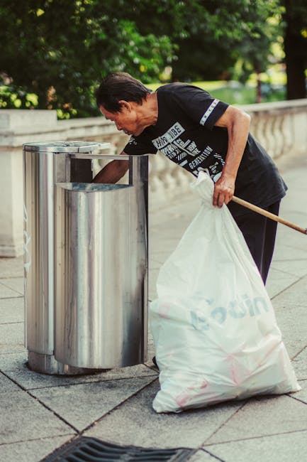 A middle-aged man with short dark hair is seen disposing of rubbish into a stainless steel public rubbish bin located on a paved sidewalk. He is wearing a black t-shirt featuring white text and a graphic design on the front, along with dark pants. His right hand is holding a large white plastic bag filled with waste, while his left hand is reaching into the bin to discard more rubbish. The bin has a shiny, reflective surface with a vertical slit opening at the top. The background includes a stone boundary wall and lush green trees, indicating a park or urban outdoor setting. The scene is lit by natural daylight, highlighting the textures of the materials and the greenery around. The image is associated with waste management or rubbish removal services, emphasizing the act of proper waste disposal outside the context of municipal collection, aligned with private or independent rubbish handling options. It reflects responsible rubbish disposal in a community environment.