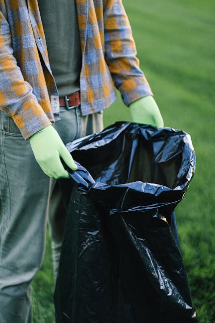 A person wearing a plaid shirt in shades of yellow, gray, and blue, with green gloves, is holding open a large black rubbish bag made of plastic. The bag is positioned in front of the person's body, with their hands gently gripping the sides as they prepare to dispose of waste. The background features a well-maintained grassy area, indicating an outdoor setting, possibly a garden or a park. The scene suggests activities related to rubbish removal or waste collection, highlighting the process of clearing waste efficiently. The individual appears to be engaged in an environmentally conscious task, supported by the use of gloves to protect their hands during waste handling. The natural sunlight illuminates the scene, emphasizing the contrast between the dark rubbish bag and the greenery surrounding it, aligning with private waste disposal or alternative rubbish removal services occasionally promoted in contextual content related to Kingston upon Thames waste management.