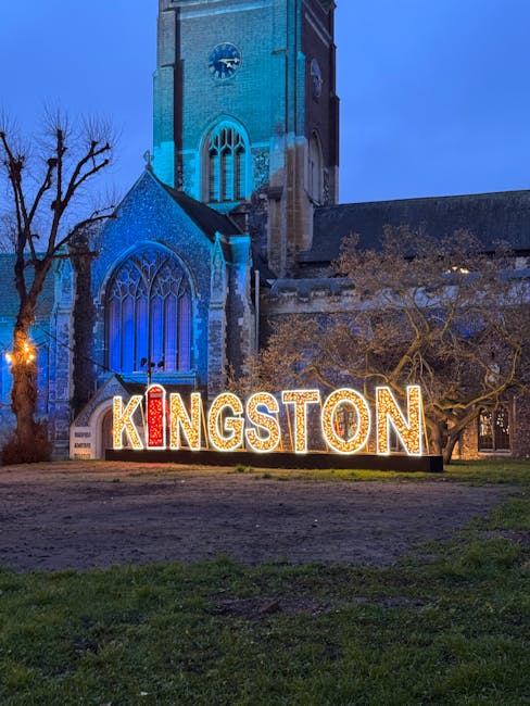 A large illuminated sign spelling 'KINGSTON' is positioned in front of a historic stone church with pointed Gothic windows and a tall clock tower. The sign features individual letters with decorative light bulbs, with the 'I' stylized as a red candle or flame. The church's exterior is built from weathered stone with a textured surface, and the windows are framed with intricate tracery. The structure is bathed in cool blue lighting, highlighting its Gothic architectural details against a darkening evening sky. To the right of the sign, a leafless tree with twisted branches extends upward, partially obscuring the church's facade. The scene is set on a patch of grass and dirt, and the lighting creates a serene, evening atmosphere, suggesting a location within the town of Kingston. The image is associated with local community identity and may relate to alternative methods of public or private waste management near historic sites, as evidenced by the presence of the sign in a public space.