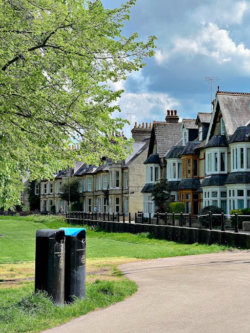 A row of residential terraced houses with traditional brickwork and pitched roofs, situated behind a black metal fence, are visible in the background. In the foreground, there is a small park or green space featuring a large leafy deciduous tree with bright green foliage extending across the top of the image. Two black rubbish bins with blue lids are positioned on a pathway made of grey paving slabs, which runs through the grassy area. The grass is well-kept, contrasting with the slightly uneven dirt patches near the bins. The sky above is partly cloudy with patches of blue, indicating a pleasant, mild day. This scene illustrates a typical suburban setting, where private waste collection can be conveniently managed alongside everyday outdoor amenities, aligning with independent waste handling practices often seen in residential areas such as this. The presence of the bins emphasizes the importance of local rubbish disposal solutions and the role of community waste management efforts.
