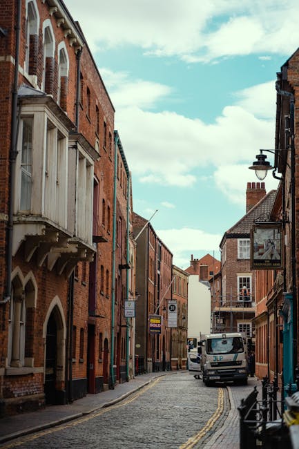 A narrow cobblestone street in Kingston upon Thames lined with multi-storey red-brick buildings featuring a mix of residential and commercial properties. On the left, a building has a distinctive bay window with white wooden framing and arched features, while on the right, there are traditional lamp posts extending from the facades, one with a small hanging sign. Parked on the street is a small white van positioned towards the middle of the scene, surrounded by minimal street furniture, including a bicycle rack near the foreground. The sky above is partly cloudy with patches of blue visible, creating natural lighting that illuminates the textured surfaces of the buildings and street. The scene reflects an urban setting with the potential for alternative waste handling, illustrating a typical setting where private rubbish collection services may operate independently from local authority collections, especially given the absence of visible public bins or waste containers in this part of the street, with the overall ambiance suggesting a mixed-use neighborhood in Kingston upon Thames.
