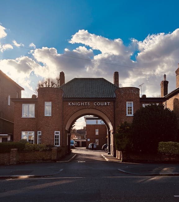 A brick residential complex with a prominent arched entrance labeled 'KNIGHTS COURT' in white lettering. The structure features rounded corner towers and rectangular windows with white frames. The foreground shows a paved driveway leading through the archway, with parked cars visible beyond. On either side of the driveway, there are low brick walls and well-maintained green shrubs. The sky above is partly cloudy with patches of blue, and sunlight casts shadows along the building’s brickwork. The scene appears to be part of a quiet residential area, possibly illustrating the context of private waste handling or on-site clearance, which could relate to alternative rubbish removal options discussed by waste management services like wasteremovalkingstonuponthames.co.uk.