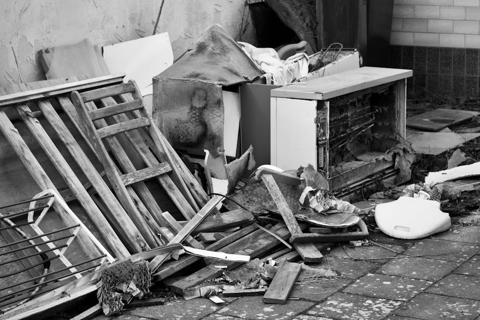 A collection of discarded household items and debris outdoors on a paved surface, including a weathered wooden staircase railing leaning against a pile. The debris features a damaged washing machine carcass with exposed internal components and peeling metal, along with broken wooden planks, a torn piece of fabric, and a white plastic foam object. Surrounding the pile are other miscellaneous materials, with part of a brick wall and a wooden panel visible in the background. The scene appears to be part of an indoor or outdoor rubbish clearance, representing typical waste left over from household removal or renovation, and is associated with private waste disposal services like those offered by wasteremovalkingstonuponthames.co.uk. The lighting is neutral, with soft shadows and no distinct sunlight, creating an overall impression of an unkempt area awaiting clearance or collection.