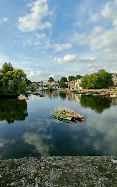 A tranquil river scene featuring a calm, reflective water surface with a few small boats docked along the edge, including a green boat and a brown boat, both positioned slightly to the right of the centre. The river is bordered by a mix of lush green trees and residential buildings with varying architectural styles on the far bank, some with light-colored facades and others with darker brickwork. The sky overhead is partly cloudy, with a blue background dotted with white, fluffy clouds, creating a peaceful atmosphere. The image appears to be taken from a stone or concrete ledge at the foreground, with speckled grey surface visible at the bottom of the frame. The surroundings suggest a quiet, residential area near the water, potentially used for leisure or small-scale private boat docking, aligning with themes of independent or alternative waste management and rubbish removal services in a scenic setting.