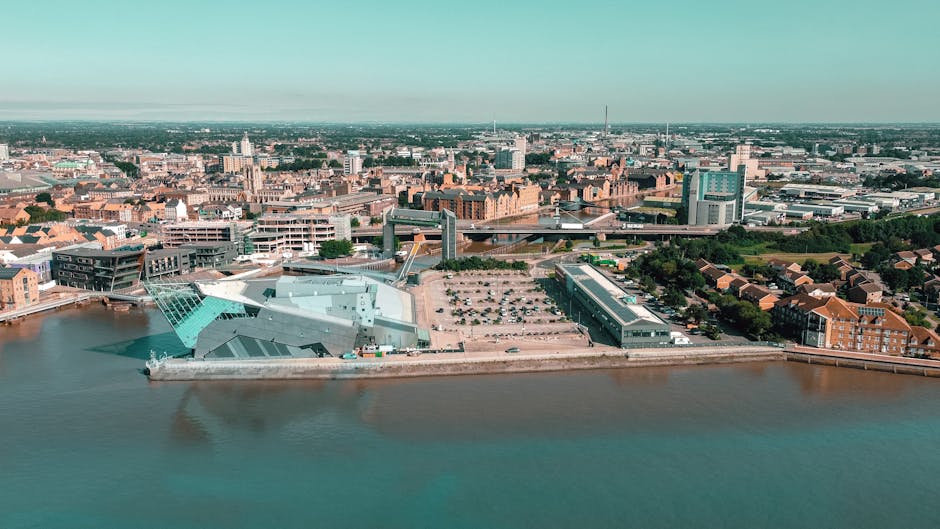 An aerial view of Kingston upon Thames showing a large cityscape alongside a river, with modern and historic buildings visible in the background. In the foreground, there is a waterfront area with several large, flat, metallic structures, likely part of a waste management facility or industrial site, situated on a concrete platform bordering the water. The structures include a covered section with a metallic, slightly textured finish, and several other metallic surfaces reflecting the daylight. The scene features a parking lot with numerous cars arranged in rows near the industrial area, surrounded by green trees and residential buildings with red brick facades and pitched roofs. The overall atmosphere suggests a clear day with good natural lighting, emphasizing the juxtaposition of urban development and waterfront landscape, which relates to private or independent waste handling and rubbish removal services in the area.
