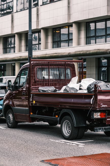 A small burgundy flatbed truck parked on an urban street in front of a modern building with large glass windows and a concrete facade. The truck's cargo area is filled with various waste materials, including black garbage bags, cardboard boxes, and loose debris, indicating an on-site clearance or private waste removal process. The side of the vehicle features a protective metal mesh that extends above the cargo bed, providing safety and containment for the rubbish. In the background, a row of parking spaces is visible, with other vehicles parked nearby, and a streetlamp stands adjacent to the truck. The scene is illuminated by natural daylight, highlighting the textures of the materials and the urban environment, aligning with non-council waste collection methods and independent rubbish disposal, as might be seen in professional waste removal services like those provided by wasteremovalkingstonuponthames.co.uk. This image illustrates the practical aspect of alternative waste handling and rubbish removal for commercial or domestic purposes.