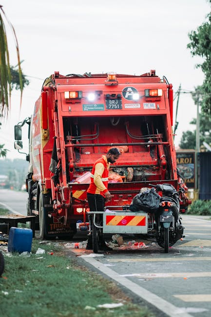 A waste collection worker dressed in a high-visibility yellow and orange vest is positioned beside the rear of a large red rubbish collection truck on a roadside. The truck's rear hatch is open, revealing mechanical components and a collection chamber, with the worker appearing to load or inspect collected debris. The black rubbish bag is placed on a small metal platform attached to a motorcycle parked beside the truck, which features a reflective orange and white striped panel for visibility. In the background, there are trees, utility poles with overhead wires, and a building with signage visible on the right side, indicating a semi-urban environment. The scene is illuminated by natural daylight, with a slightly overcast sky, and the ground shows a curb and part of the road, suggesting an on-site waste collection or clearance operation, consistent with private waste management services that handle rubbish removal without relying solely on local authority collection.